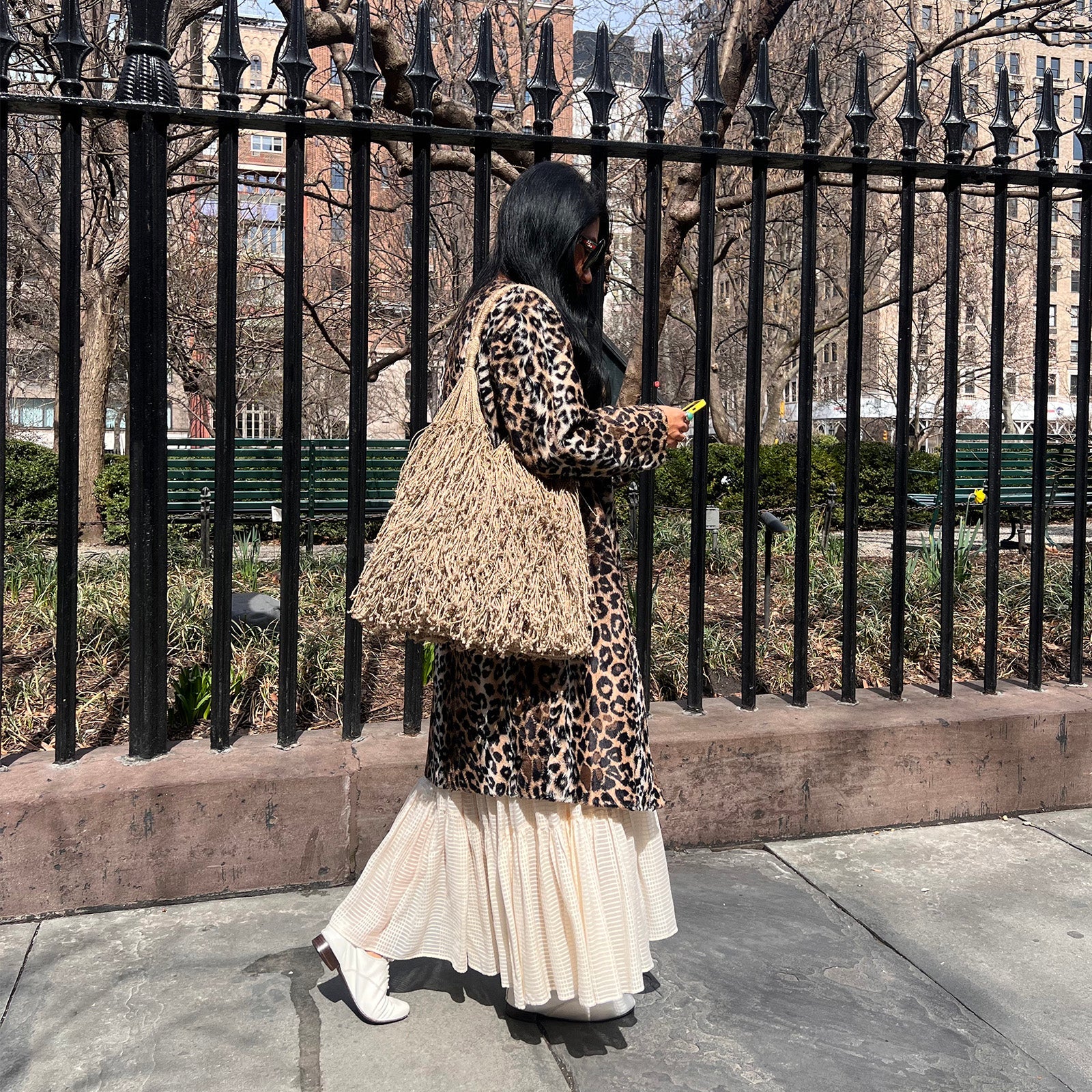 Woman in leopard coat and flowing skirt walks by a black fence. She carries a large woven bag, holding a phone, exuding a chic urban vibe.