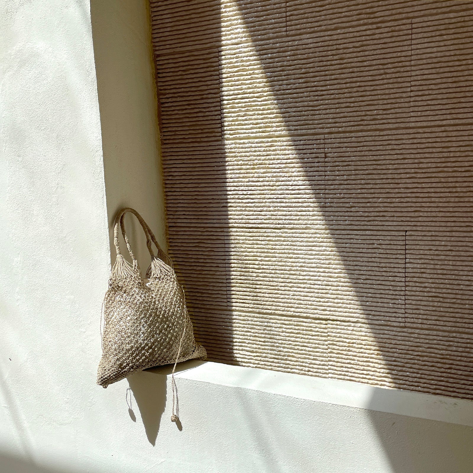A woven straw bag hangs on the edge of a white ledge, with sunlight casting shadows across a textured beige wall in the background.