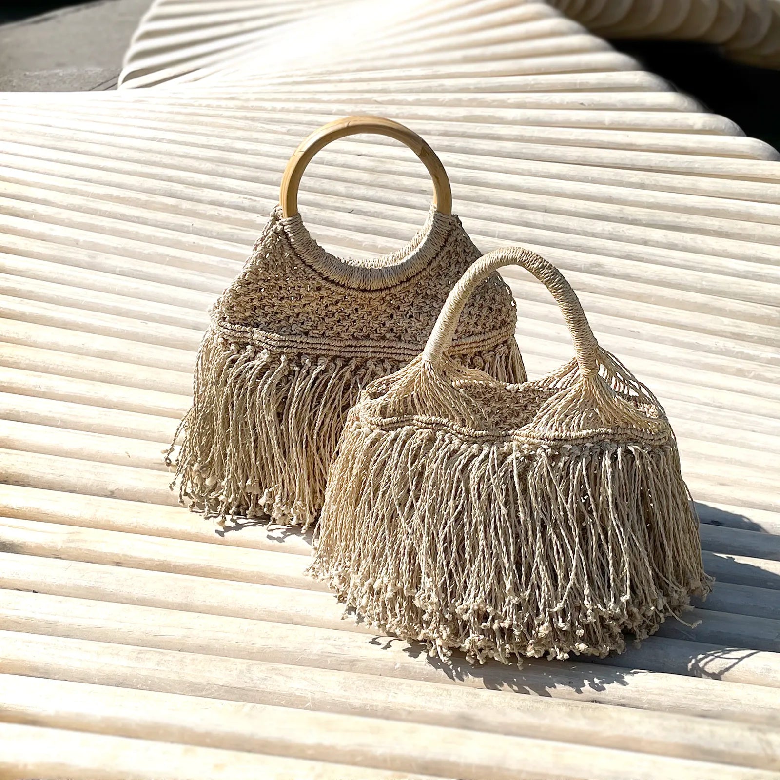 Two woven straw bags with fringe detailing are displayed on a textured, sunlit surface. One bag has a wooden handle, creating a natural, bohemian vibe.