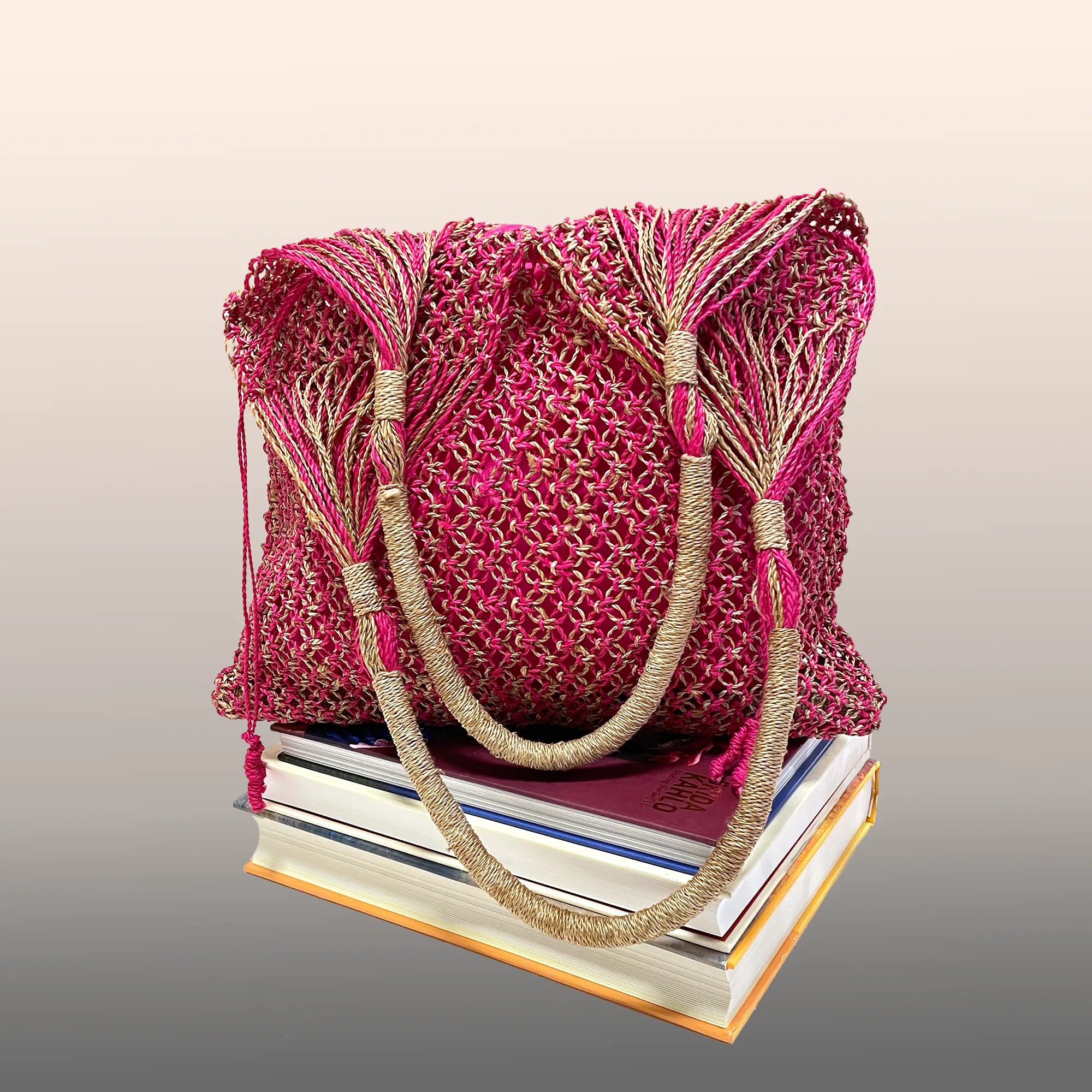 A pink and gold woven Majuun handbag with two handles rests atop a stack of three closed books against a light gradient background.