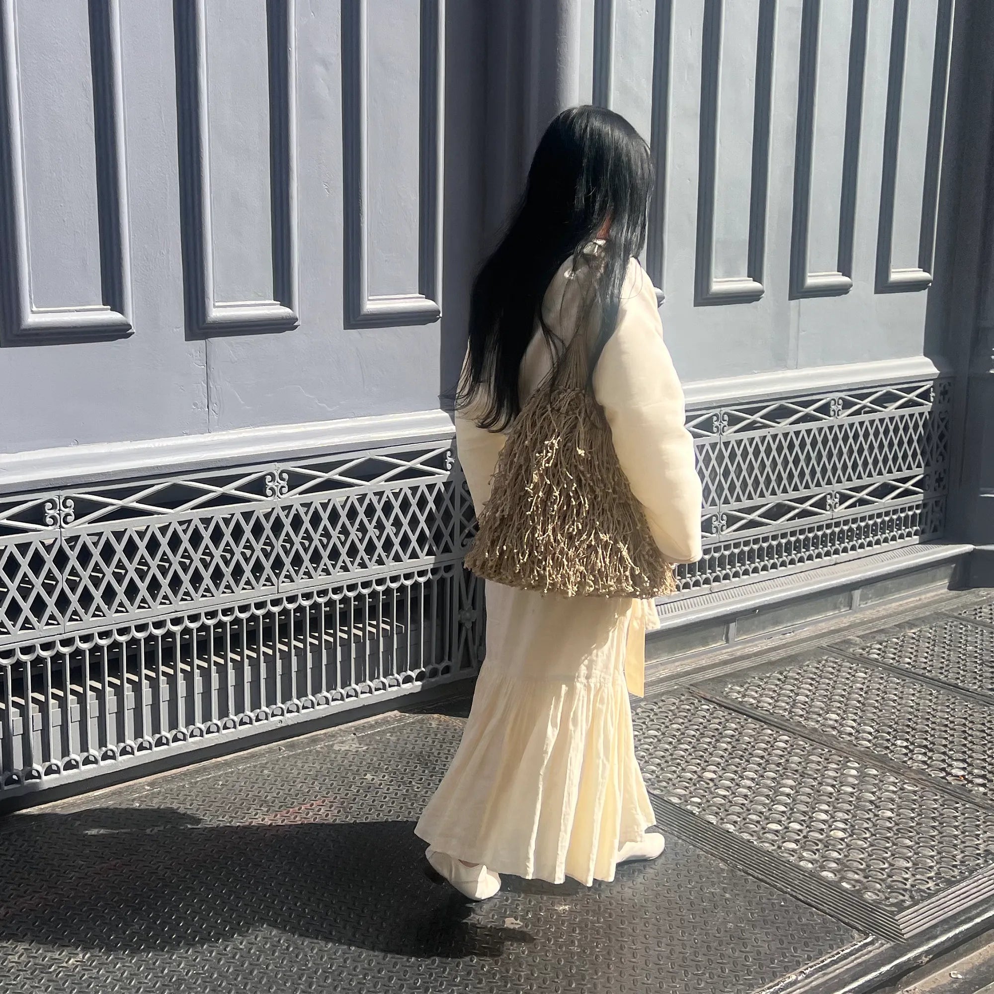 Woman in a long white dress with a textured brown bag walking on a sidewalk.
