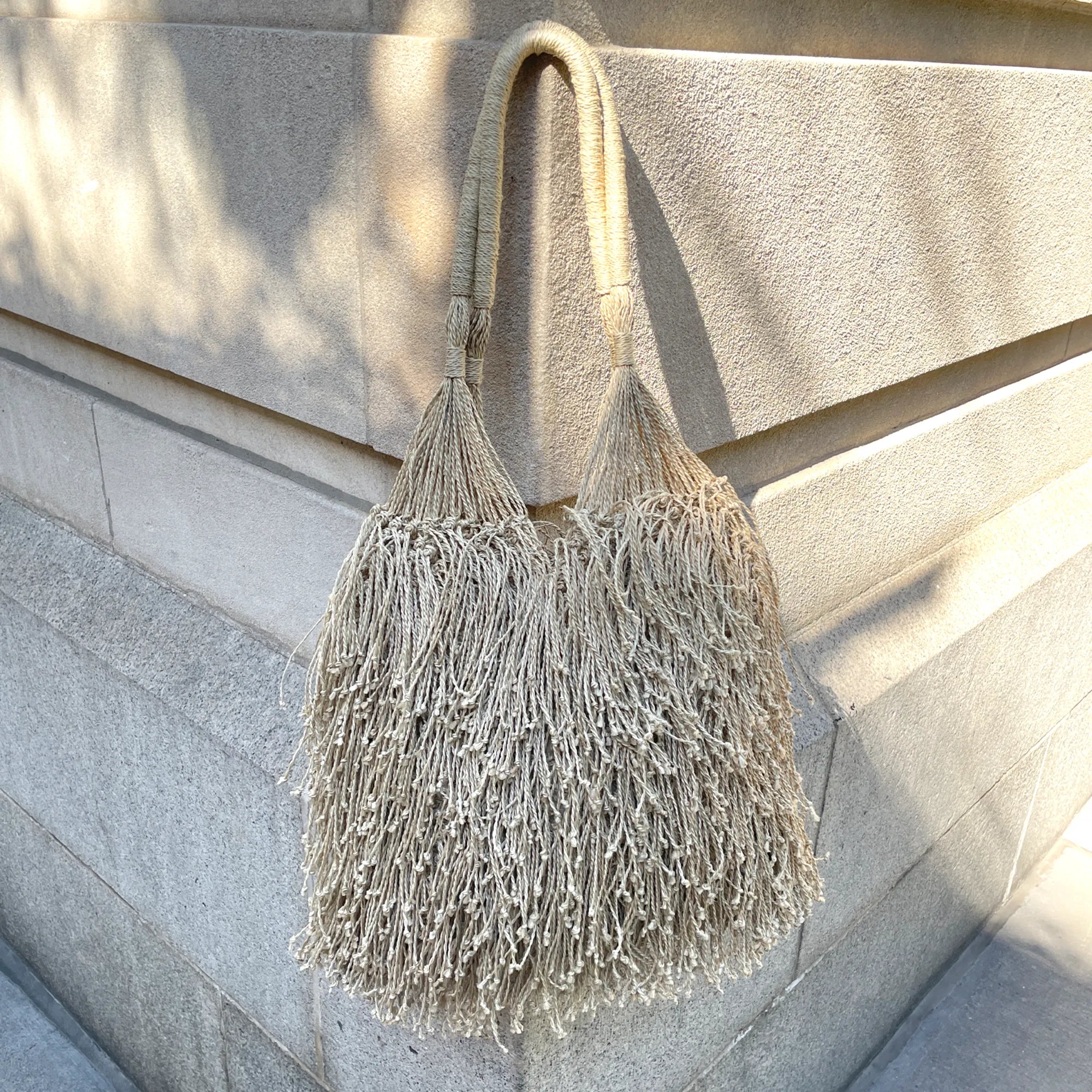 Beige handbag with fringe details against a stone wall.