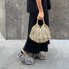 Person holding a woven handbag on a sidewalk with a gray wall background