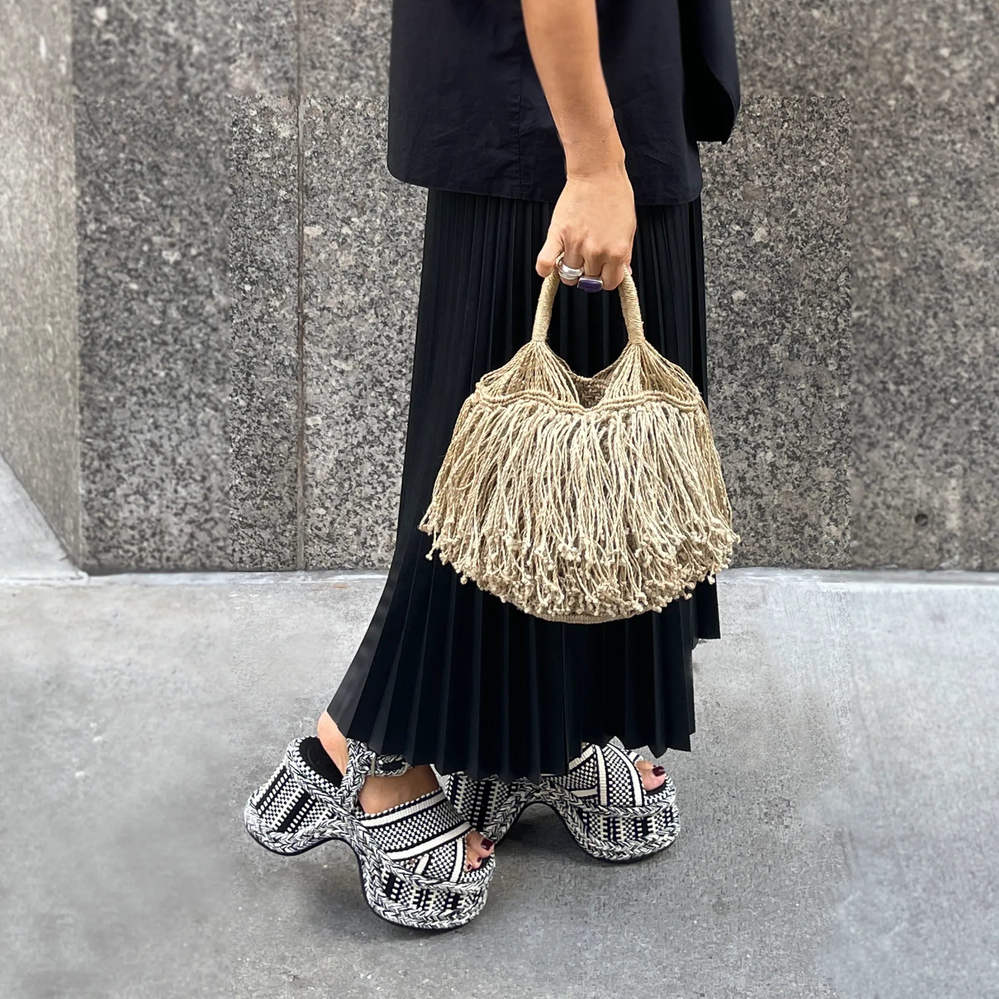 Person holding a woven handbag on a sidewalk with a gray wall background