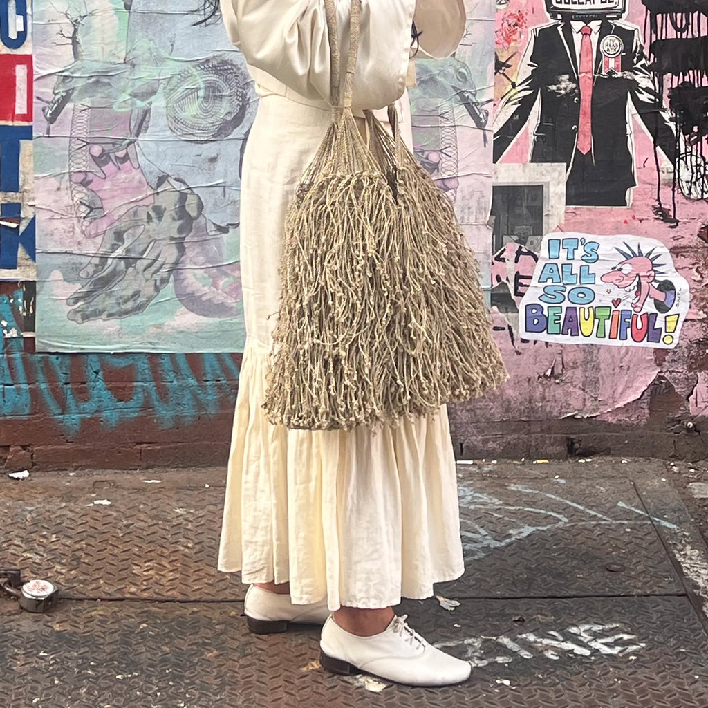 Person wearing a long white dress and holding a textured bag against a colorful mural background.