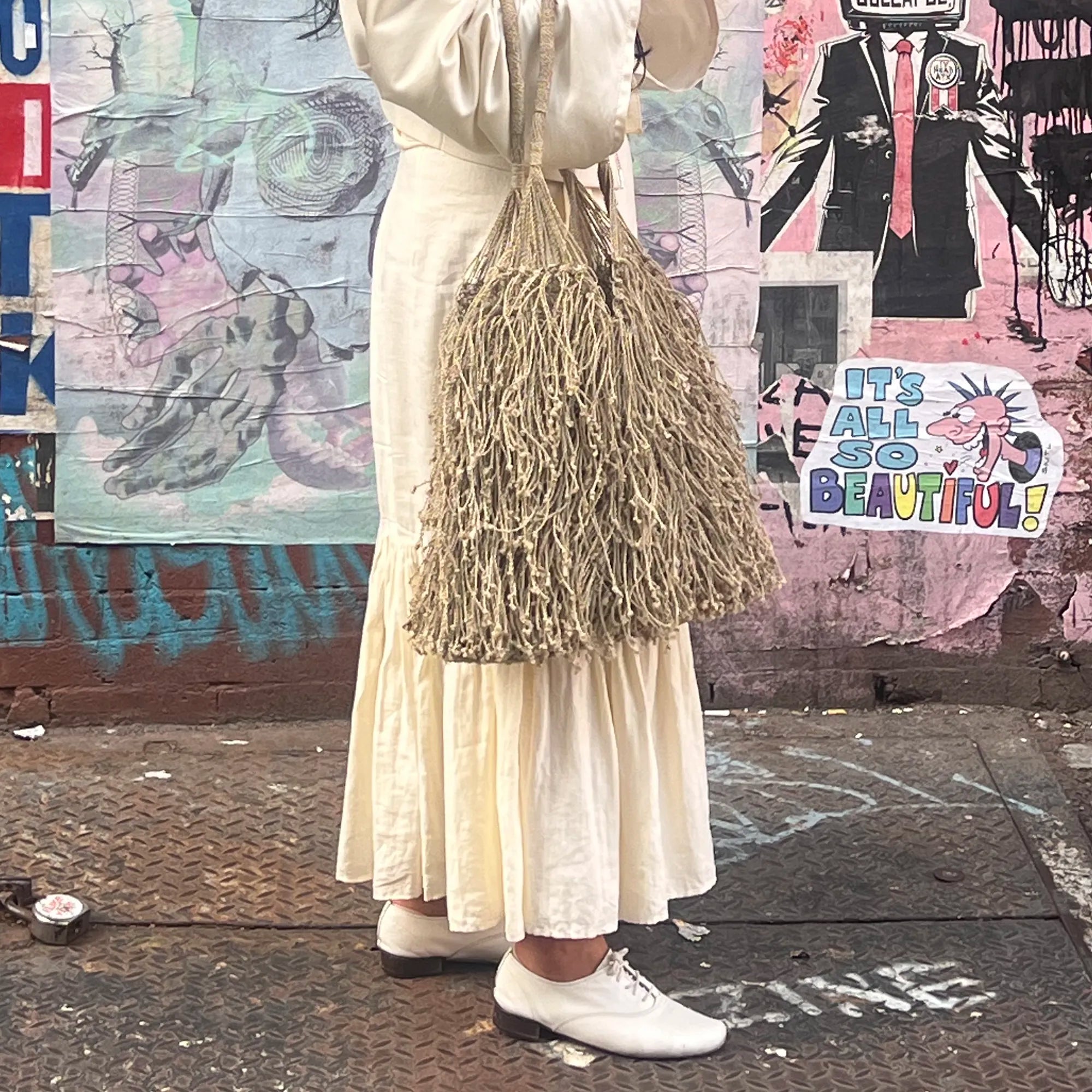 Person wearing a long white dress and holding a textured bag against a colorful mural background.