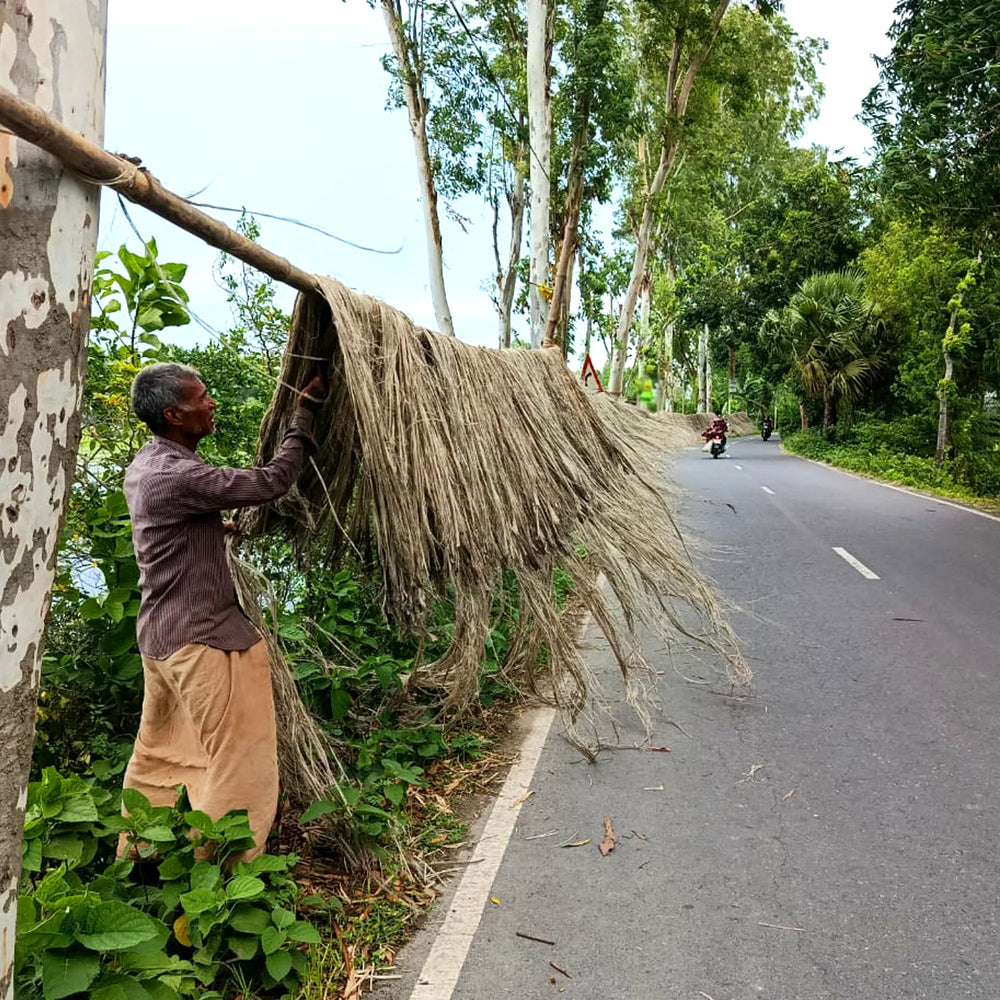 Man holding a large bundle of dried grass or straw next to a road with trees and a motorcycle in the background.